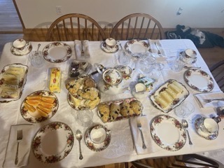 A picture of a tea party setup with a teapot, cups, and plates of food on a table with a floral tablecloth.